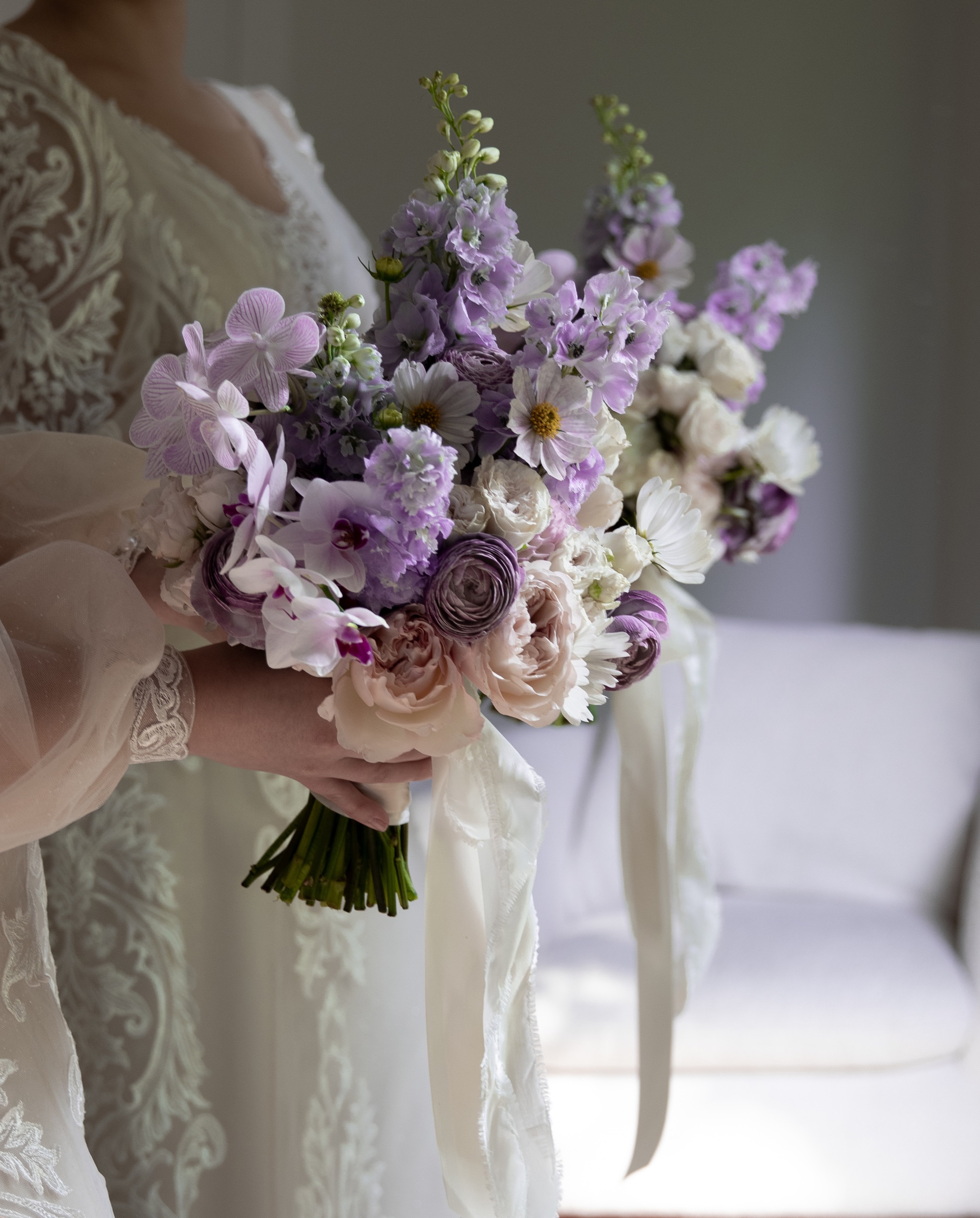 A bride in lace holding a lavender wedding bouquet in soft light.