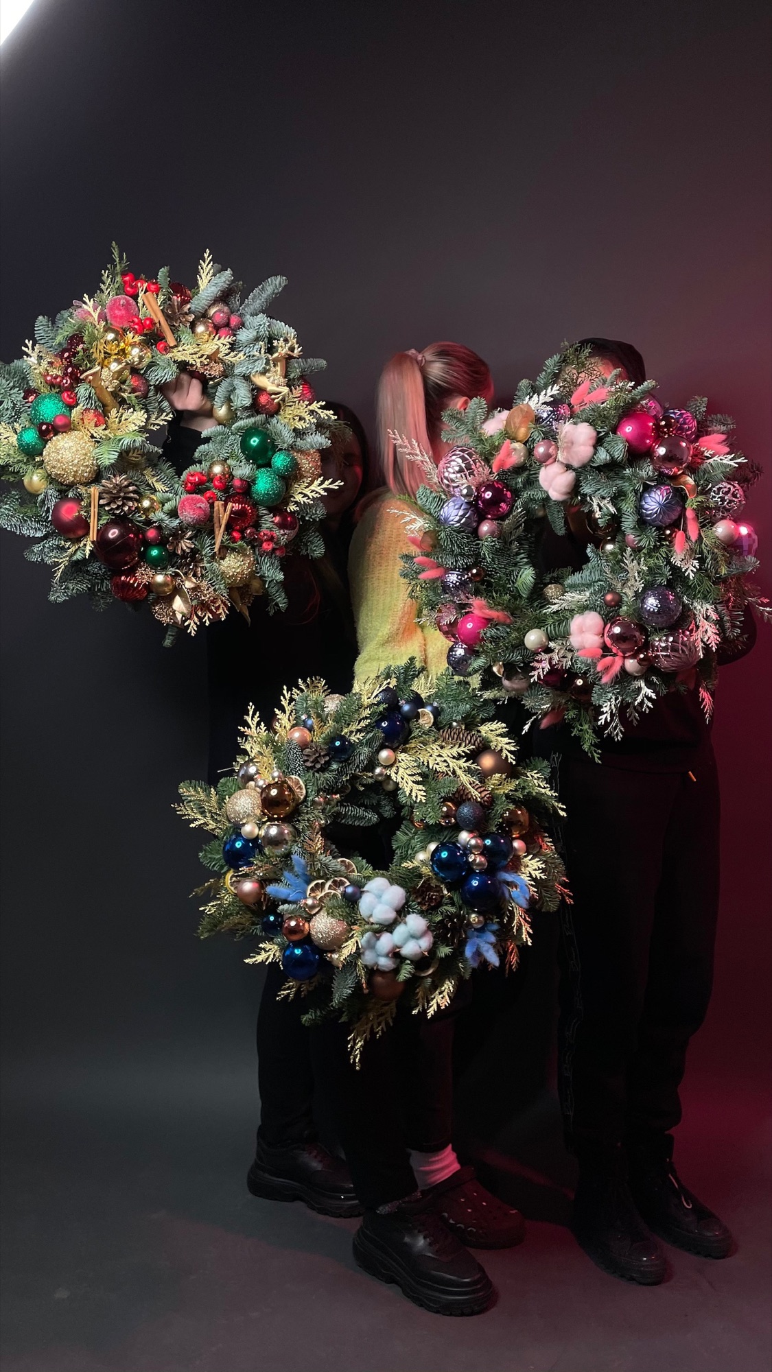 Two florists holding three ornate Christmas wreaths on a dark backdrop