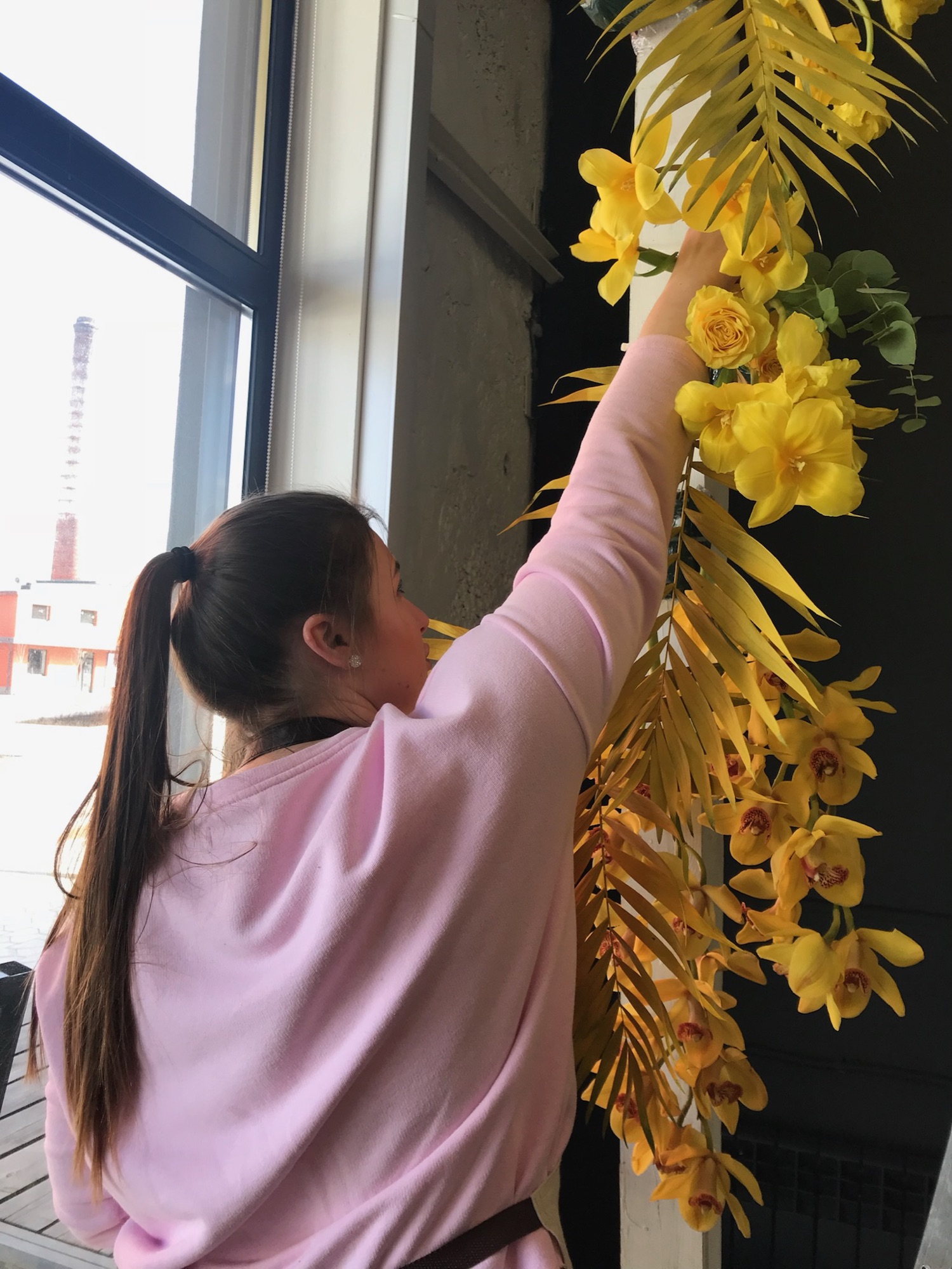 A florist reaching up to install a yellow orchid garland by a studio window
