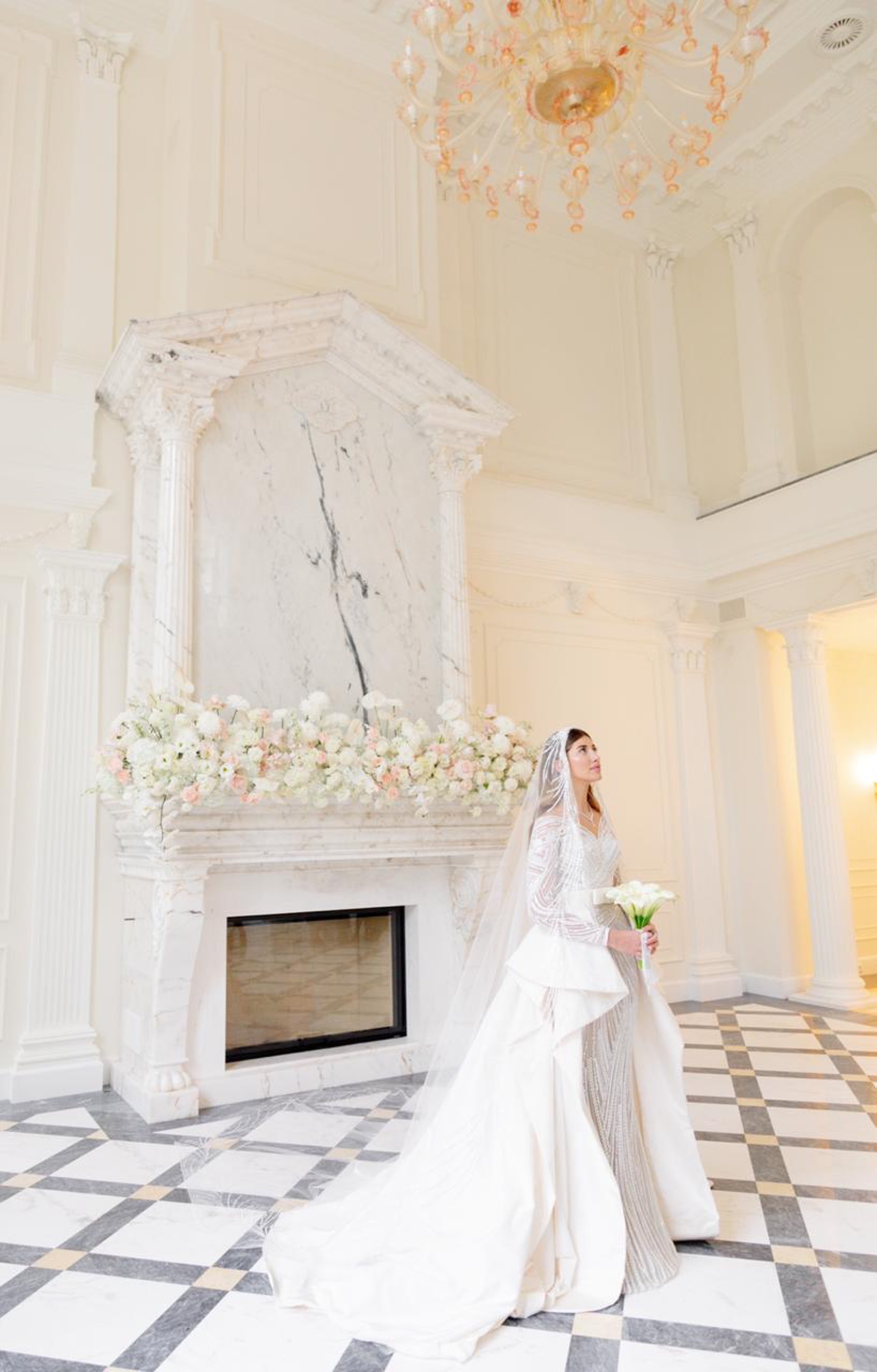 A bride under a chandelier beside a floral mantel installation in a grand hall